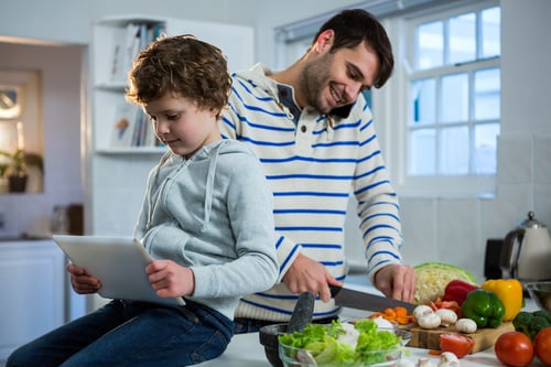Father talking on mobile phone while chopping vegetables and son using digital tablet in the kitchen